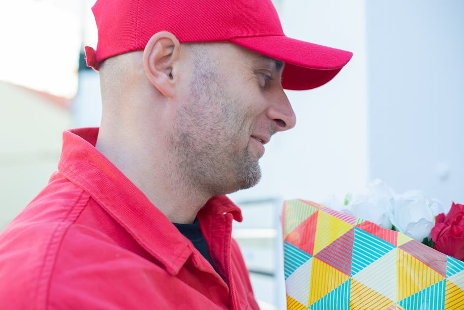 A delivery man in a red uniform and matching red cap holding a colorful, geometric-patterned gift bag filled with white and red flowers. The man is smiling gently and looking at the floral arrangement