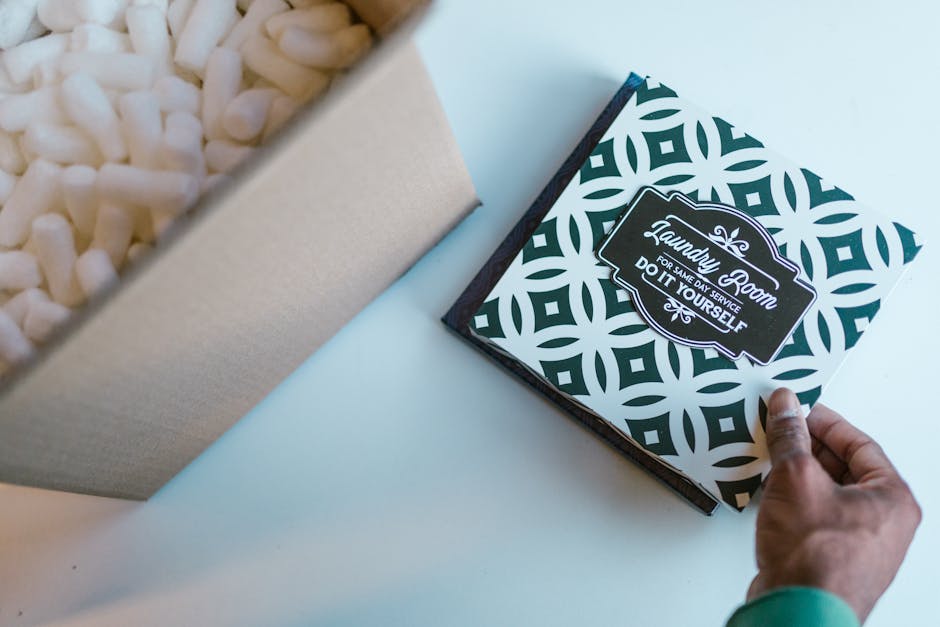 A person's hand opening a black and white patterned box labeled 'Laundry Room' with the subtitle 'For Same Day Service Do It Yourself', next to an open beige box filled with white packing material, on