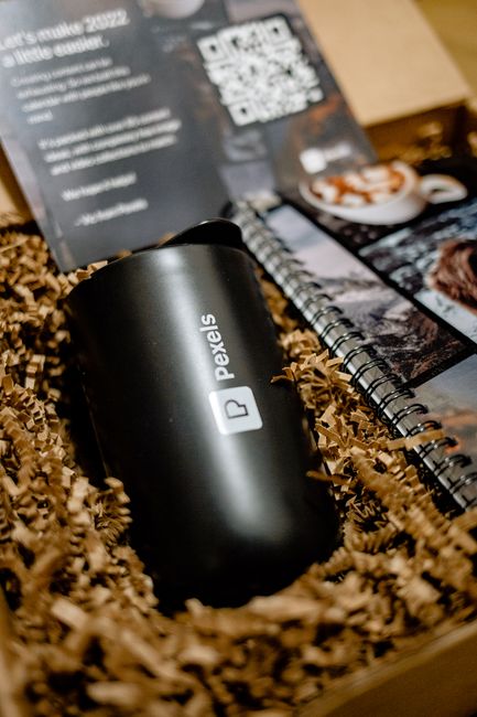 A sleek black insulated tumbler with the Pexels logo printed on it, placed among crinkled brown packing paper inside a cardboard box. In the background, there is a black and white printed card with a 
