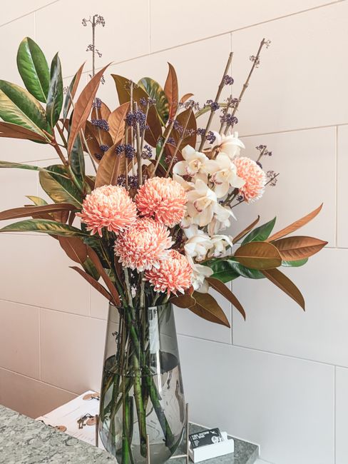 A tall, clear glass vase filled with a fresh floral arrangement featuring large, soft pink dahlias, white orchids with light pink accents, and sprigs of lavender. The bouquet includes broad, dark gree