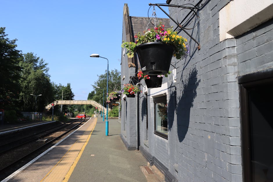 A street scene at Harold Wood station with a focus on hanging flower baskets attached to a gray brick building. The baskets contain colorful blooming flowers including pink, yellow, purple, and white