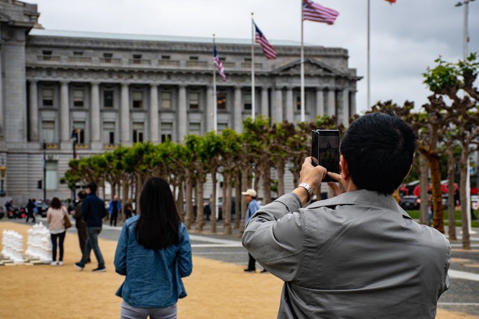 A person with short black hair, wearing a light gray jacket, is holding a smartphone in both hands and taking a photo of the US Capitol building in Washington, D.C., during daytime. The building's neo