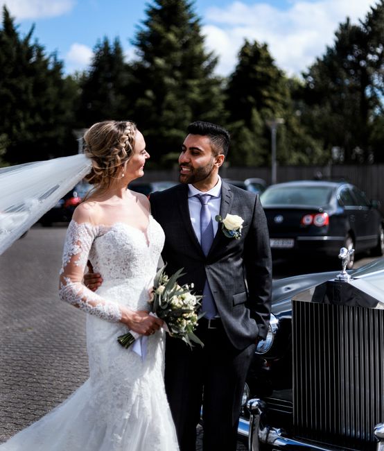 A bride and groom sharing a joyful moment outdoors, with the bride in an elegant white lace wedding gown featuring long sleeves and a delicate veil, holding a bouquet of white roses and greenery. The 