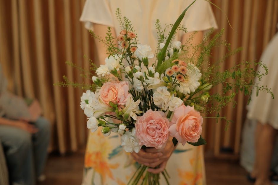 A person holding a large floral bouquet featuring soft pink roses, white lisianthus, small peach-colored daisy-like flowers, and delicate green foliage arranged in a loose, natural style. The bouquet 
