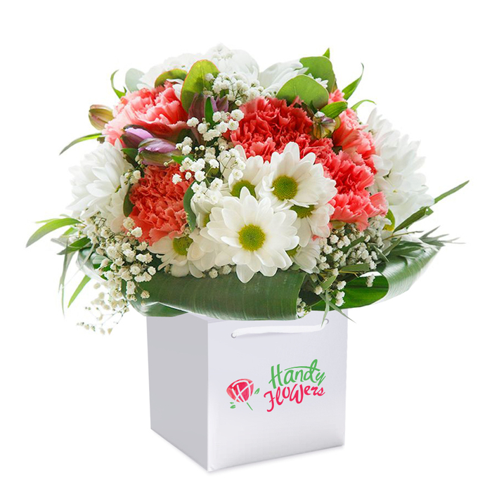 Standing elegantly in a pristine square white gift bag, this floral bouquet showcases the skill and nuance that Harold Park's finest florists bring to every creation. The eye is immediately drawn to the full, rounded profusion of soft coral and radiant red carnations, their frilled petals layered generously for a texture that feels both abundant and plush. Dancing among these focal blooms are crisp white daisies, their zestful lime-green cores catching the light much like wildflowers growing in the open meadows near Harold Court Woods. Wisps of delicate white baby's breath thread throughout, adding gentle airiness that hints at the lightest of breezes. Along the bouquet's left edge, a few unopened, purple-brushed buds introduce a sense of mystery and anticipation, subtly enhancing the depth of the arrangement. Broad, vibrant green foliage envelopes the whole, fanning out in glossy arcs and offering a lush, living cradle at the base. The composition is seamless, reflecting a careful balance and a deep understanding of colour and form. Presented in a clean, minimalist white bag with sturdy ribbon handles, the bouquet exudes readiness for gifting-whether as a celebratory centrepiece for a milestone at Central Park Community Hub, a cheerful birthday surprise along Gubbins Lane, or a comforting gesture of thoughtfulness for a Harold Park neighbour. Every element speaks to freshness, artistry, and the joy of thoughtfully delivered flowers in this close-knit local community.